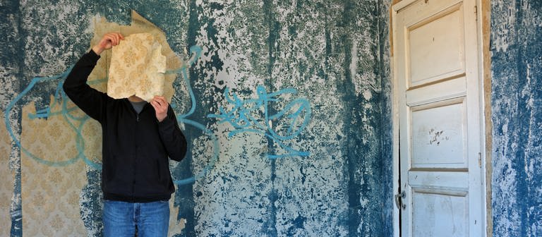 Psychologie. Ein Mann hält sich in einem blauen Raum ein Stück Tapete vors Gesicht (Foto: Colourbox)