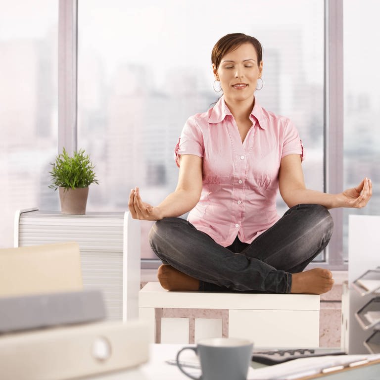 Eine Frau sitzt meditierend auf ihrem Schreibtisch: Stressbewältigung im Job ist wichtig für die eigene Gesundheit (Foto: IMAGO, IMAGO / Shotshop)
