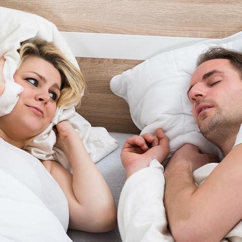 Eine Frau hält sich im Bett mit einem Kissen die Ohren zu und schaut ihren schnarchenden Mann an. (Foto: Getty Images, Thinkstock -)