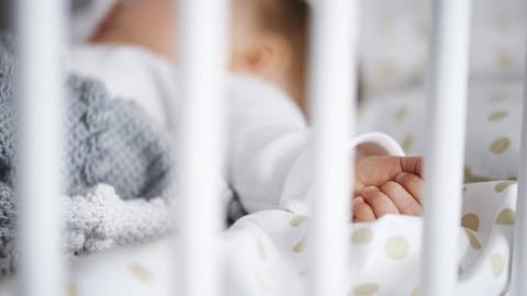 Ein Baby schläft in Rückenlage im Gitterbett (Foto: IMAGO, imago/Westend61)