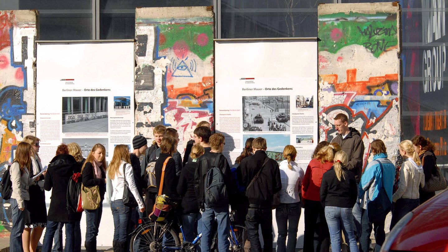 imago/PEMAX (Foto: IMAGO, Schüler während einer Geschichtsstunde vor Resten der Berliner Mauer und Informationstafeln am Potsdamer Platz in Berlin (2007))