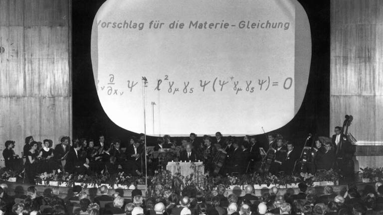 Bei einem Festakt zum 100. Geburtstag von Max Planck (23. Januar 1958) am 25. April 1958 in der Westberliner Kongreßhalle erläutert Werner Heisenberg (am Rednerpult) seine vieldiskutierte "Weltformel", die er am 28. Februar 1958 erstmals vorgestellt hatte: Die Theorie der kleinsten Teile verbindet die allgemeine Relativitätstheorie mit der Quantentheorie zu einer allgemeinen Feldtheorie.  (Foto: picture-alliance / Reportdienste, picture-alliance / dpa | Günter Bratke)