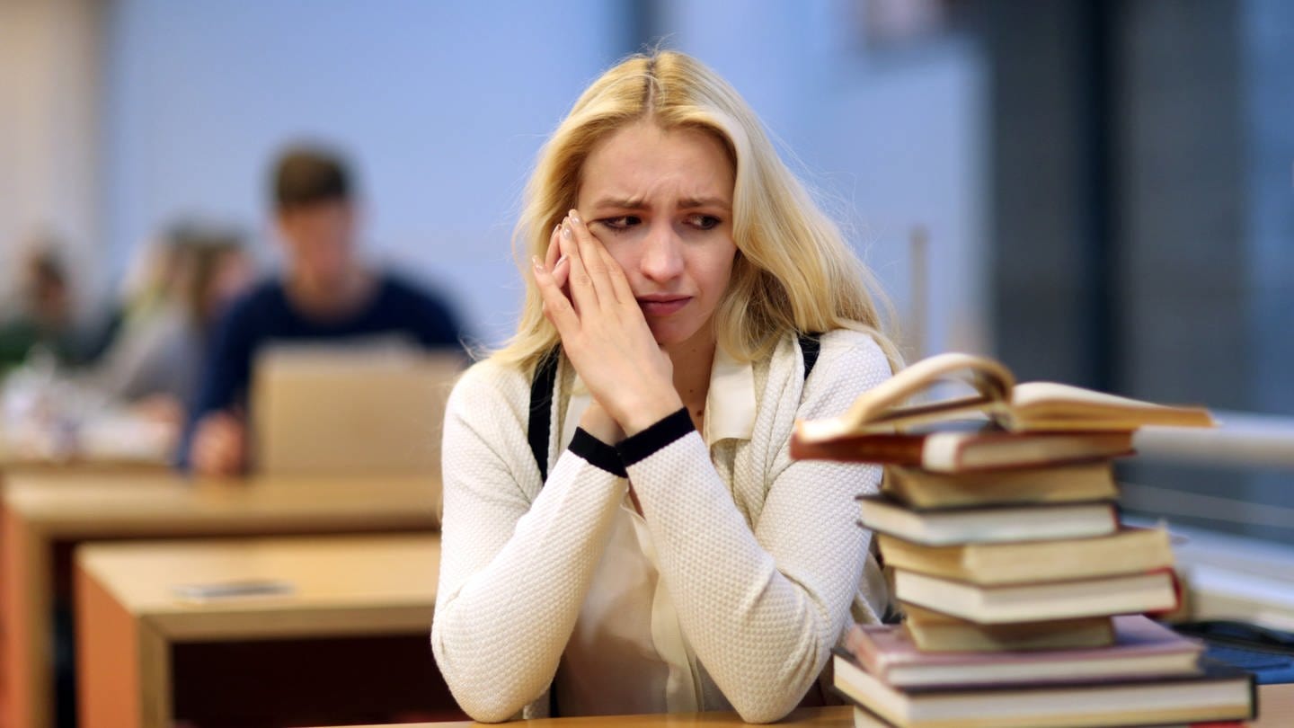 Eine junge Frau sitzt in einer Bibliothek und starrt auf einen Bücherstapel: Vor allem wer selbstständig am Computer arbeitet oder studiert, ist Prokrastination bzw. Aufschieberitis konfrontiert