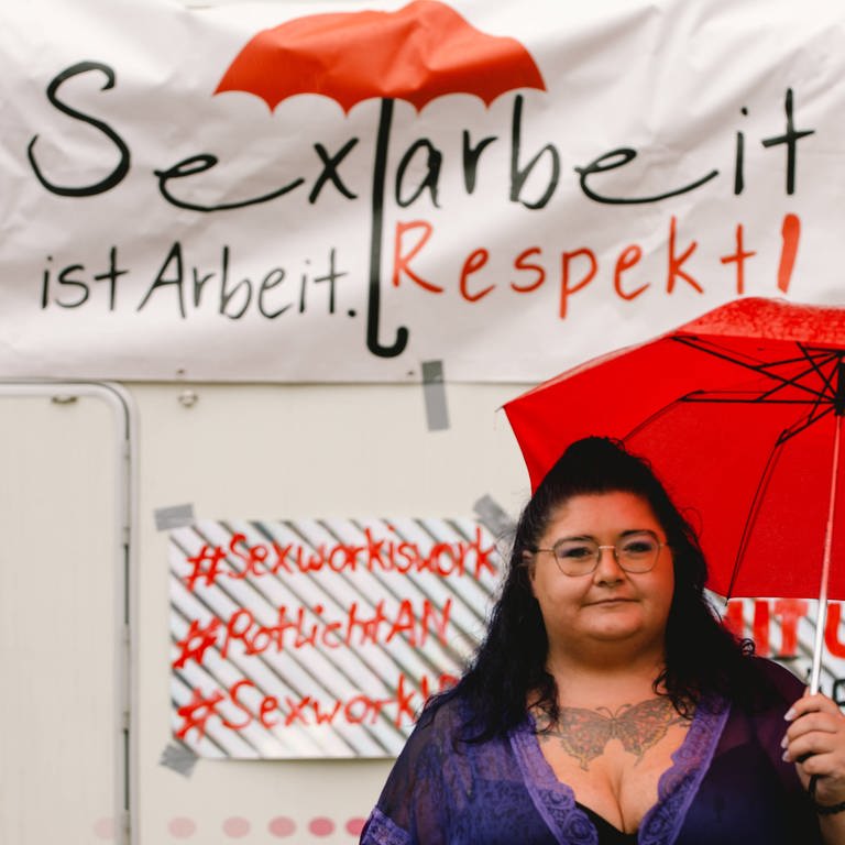 Eine Sexarbeiterin mit rotem Regenschirm in der Hand demonstriet am 22. Juni 2021 vor dem Kölner Dom vor einem Schild mit der Aufschrift: "Sexarbeit ist Arbeit. Respekt!" (Foto: IMAGO, IMAGO / NurPhoto)