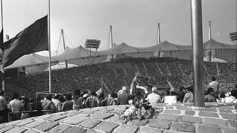Blumen liegen am 6. September 1972 während des Gedenkgottesdienstes für die elf ermordeten israelischen Teammitglieder am Fuße der olympischen Fackel im Münchner Olympiastadion, eine Fahne weht auf Halbmast. Das Stadion mit 80.000 Sitzplätzen war während der Zeremonie bis auf den letzten Platz gefüllt. (Foto: picture-alliance / Reportdienste, picture alliance / AP | AP)