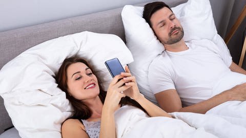 Symbolfoto: Eine Frau benutzt ihr Smartphone im Bett während der Ehemann schläft (Foto: IMAGO, IMAGO / Panthermedia)