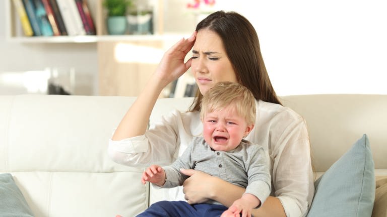 Gestresste Mutter hält weinendes Kleinkind auf dem Schoß: Perfekt organisiert und gelassen, vormittags erfolgreich im Beruf und nachmittags Spielkameradin und Hausaufgabenbetreuerin. Die Anforderungen an eine "gute Mutter" sind heute groß – und oft widersprüchlich. Das idealisierte Mutterbild prägt auch die hohen Ansprüche, die viele Mütter an sich selbst stellen. Der Mythos der angeborenen, bedingungslosen Mutterliebe sitzt tief.  (Foto: IMAGO, IMAGO / Panthermedia)