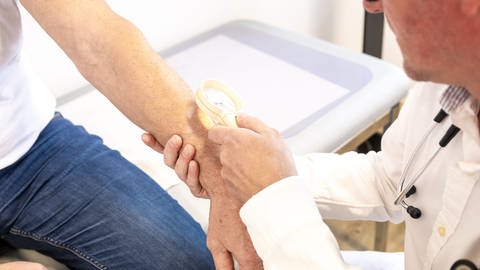 Ein Hausarzt untersucht in seiner Praxis die Haut am Arm eines Patienten mit Hilfe einer Lupe (Foto: picture-alliance / Reportdienste, picture alliance / dpa-tmn | Benjamin Nolte)