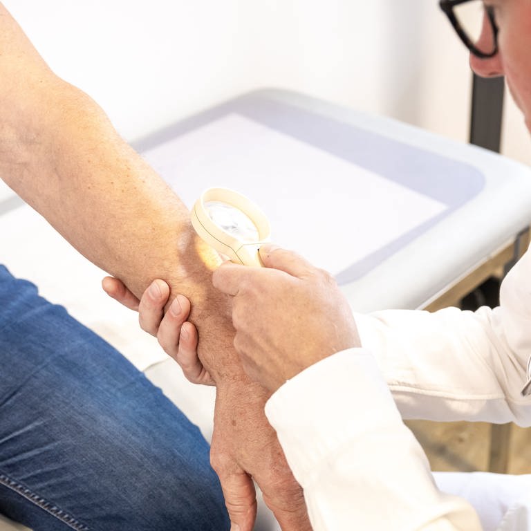 Ein Hausarzt untersucht in seiner Praxis die Haut am Arm eines Patienten mit Hilfe einer Lupe (Foto: picture-alliance / Reportdienste, picture alliance / dpa-tmn | Benjamin Nolte)