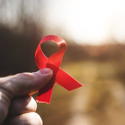 Eine Hand hält eine rote Schleife gegen die Sonne. Symbolbild für Aids und HIV.  (Foto: IMAGO, IMAGO / Bihlmayerfotografie)
