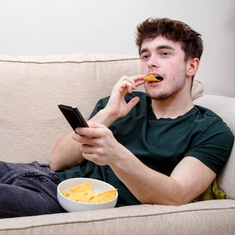 Ein junger Mann liegt auf dem Sofa und isst Chips. (Foto: IMAGO, IMAGO / Pond5 Images)