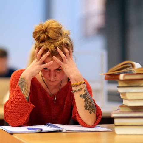 Eine Frau sitzt verzweifelt in der Bibliothek einer Uni am Tisch und stützt ihren Kopf mit beiden Händen.  (Foto: picture-alliance / Reportdienste, picture alliance / dpa Themendienst | Markus Hibbeler)