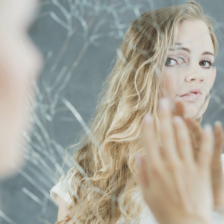 Junge Frau schaut in einen kaputten Spiegel: "Warum hat es mich erwischt?" Das fragen sich viele Kranke. Der Zusammenhang zwischen Psyche und Krankheit beschäftigt Forschende. (Foto: IMAGO, IMAGO / Pond5 Images)