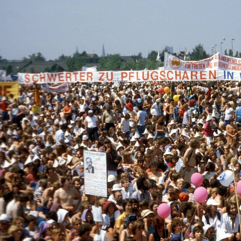 Demonstration auf den Rheinwiesen bei Bonn gegen die nukleare Nachrüstung am 10. Juni 1982. Etwa 500.000 Menschen nahmen teil. Im Hintergrund ein großes Transparent mit der Aufschrift: "Schwerter zu Pflugscharen – In West und Ost". Das Zitat "Schwerter zu Pflugscharen" hat seinen Ursprung in der Bibel und wurde auch in der Friedensbewegung zum geflügelten Wort. (Foto: IMAGO, IMAGO / Sven Simon)