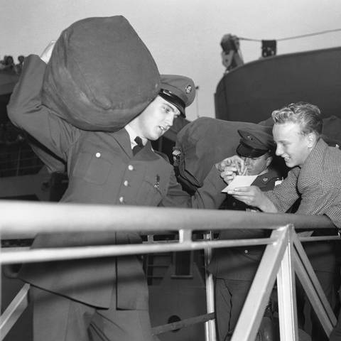 Der Rock-and-Roll-Sänger Elvis Presley gibt einem Fan ein Autogramm, während er nach seiner Ankunft in Bremerhaven am 1. Oktober 1958 sein Gepäck auf der Schulter über die Gangway schleppt (Foto: picture-alliance / Reportdienste, picture alliance / ASSOCIATED PRESS)