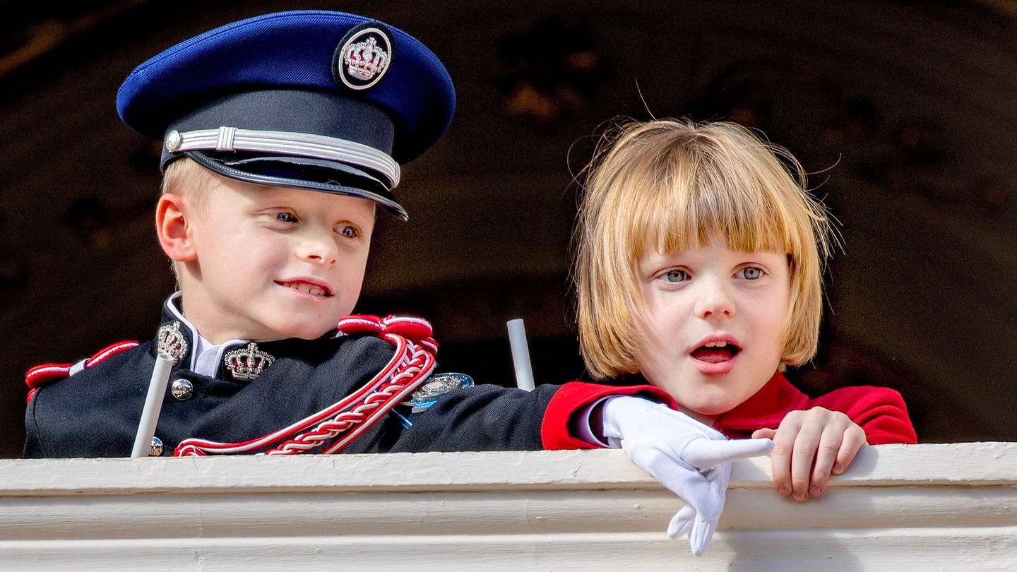 Prinz Jaques und seine Zwillingsschwester Prinzessin Gabriella von Monaco stehen auf einem Balkon, lehnen am Geländer und beobachten am Nationalfeirtag etwas mit großem Interesse.