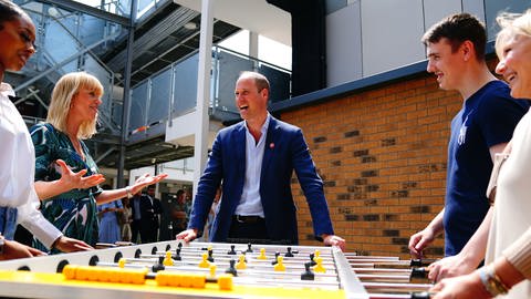 Prinz William steht im blauen Sakko und hellblauem Hemd lässig an einen gelben Tischkicker gelehnt. Die vier Spieler*innen und er lachen und haben Spaß. (Foto: dpa Bildfunk, picture alliance/dpa/PA Wire | Victoria Jones)