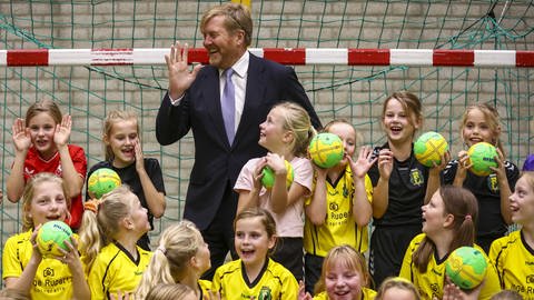 Blonder Mann mit erhobener Hand steht in einem Handballtor umringt von Mädchen in gelbem Trikot