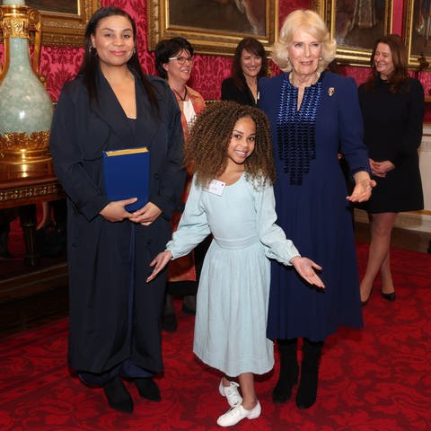 Ein kleines Mädchen mit hellblauem Kleid und stark gelockten Haaren steht vor einer dunkelhaarigen Frau und eine grauhaarigen Frau im duenkelblauen Kleid - Königin Camilla - und macht einen Knicks