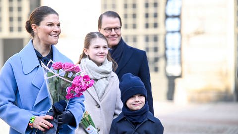 Kronprinzessin Victoria von Schweden trägt einen hellblauen Mantel und einen pinken Blumenstrauß. Neben ihr steht ihre Familie: Tochter Estelle und Sohn Oscar sowie ihr Ehemann Prinz Daniel von Schweden. Alle lächeln und sind warm gekleidet, denn sie stehen im Innenhof des Schlosses von Stockholm um ihren Namenstag zu feiern.