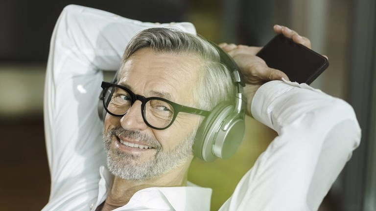 Podcast hören und abonnieren: Ein Mann mit grauen Haaren und grauem Bart hört etwas über Kopfhörer vom Smartphone und lächelt dazu.