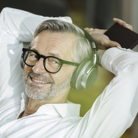 Podcast hören und abonnieren: Ein Mann mit grauen Haaren und grauem Bart hört etwas über Kopfhörer vom Smartphone und lächelt dazu.