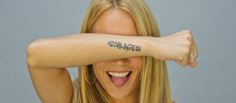 Sonia Liebing hält vor einer grauen Wand ihren mit "Schlager" tatowierten Arm vor die Augen und streckt die Zunge raus (Foto: SWR)