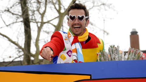 Florian Silbereisen trägt eine bunte Uniform und eine herzförmige Brille. Er Ist auf einem Karnevalswagen und jubelt der Menge zu. (Foto: picture-alliance / Reportdienste, Picture Alliance)