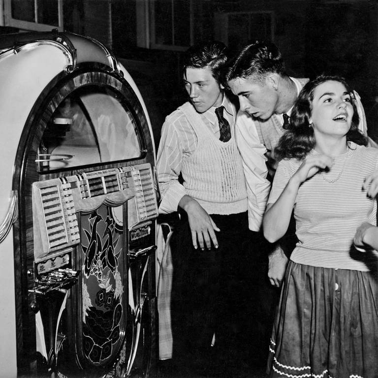 Schwarz-weiß Bild aus den 1950er-Jahren. Junge Menschen stehen vor einer Jukebox. (Foto: picture-alliance / Reportdienste, picture alliance / World History Archive | -)