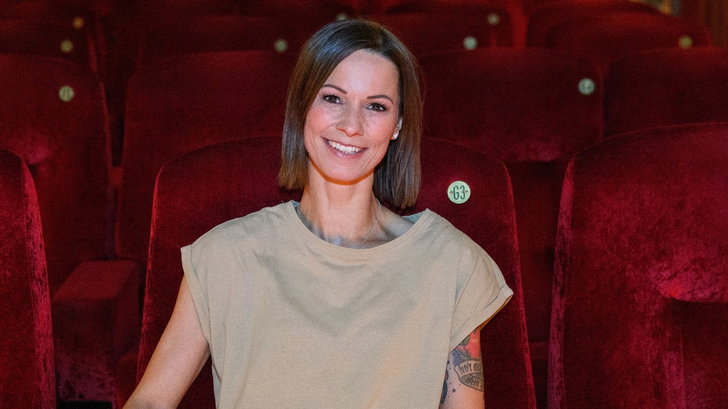 Sängerin Christina Stürmer trägt ein T-Shirt in der Farbe beige. Sie lächelt und sitzt in einem Kinosaal voller samtiger roter Sitze. An ihrem linken Arm sind Tattoos zu sehen. (Foto: picture-alliance / Reportdienste, picture alliance/dpa | Bernd von Jutrczenka)