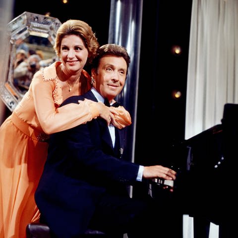 Sängerin Johanna von Koczian umarmt den Entertainer Peter Alexander von hinten, während er am Klavier sitzt. Das Bild stammt aus einer Fernsehsendung aus dem Jahr 1977.