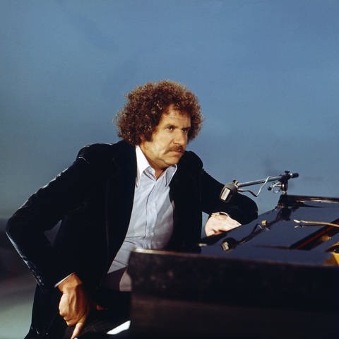 Mort Shuman sitzt an einem Piano vor einem blauen Hintergrund.