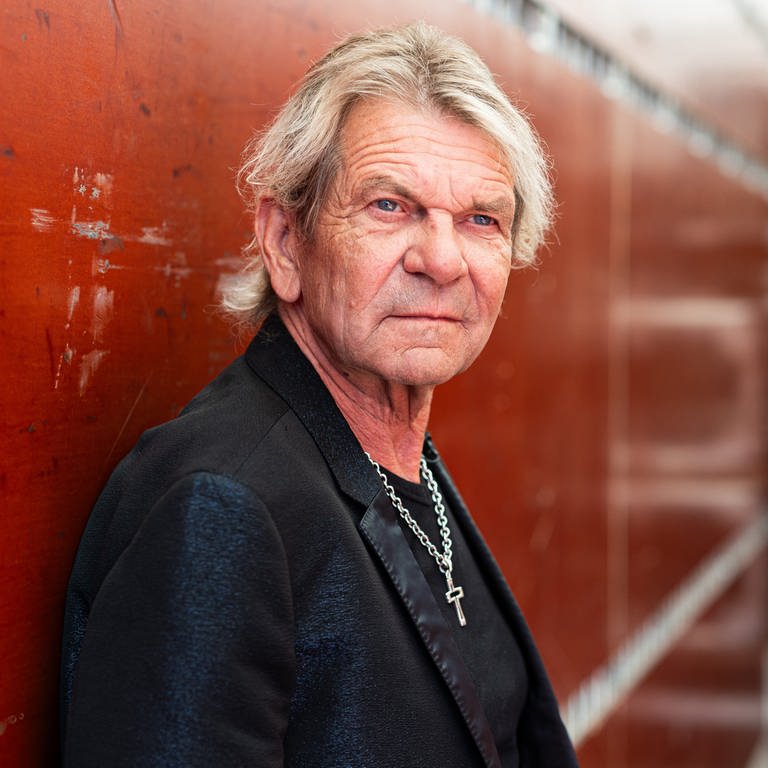 Portrait von Schlagersänger Matthias Reim in einer schwarzen Jacke vor einer roten Mauer.