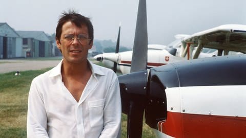 Liedermacher, Chanson-Sänger und Hobbypilot Reinhard Mey ("Über den Wolken") steht in einem kurzärmlichen Hemd neben einem Flugzeug und schaut in die Kamera. (Foto: picture-alliance / Reportdienste, picture-alliance / dpa | Ossinger)