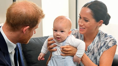 Herzogin Meghan hält ihren Sohn Prinz Archie hoch, Prinz Harry sitzt daneben und schaut zu (Foto: dpa Bildfunk, Picture Alliance)