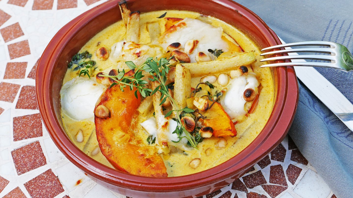 braune Tonschale, darin Kürbisspalten und Kohlrabistifte in Currysoße mit Erdnüssen und hellem Ziegenkäse, mit Käutern garniert