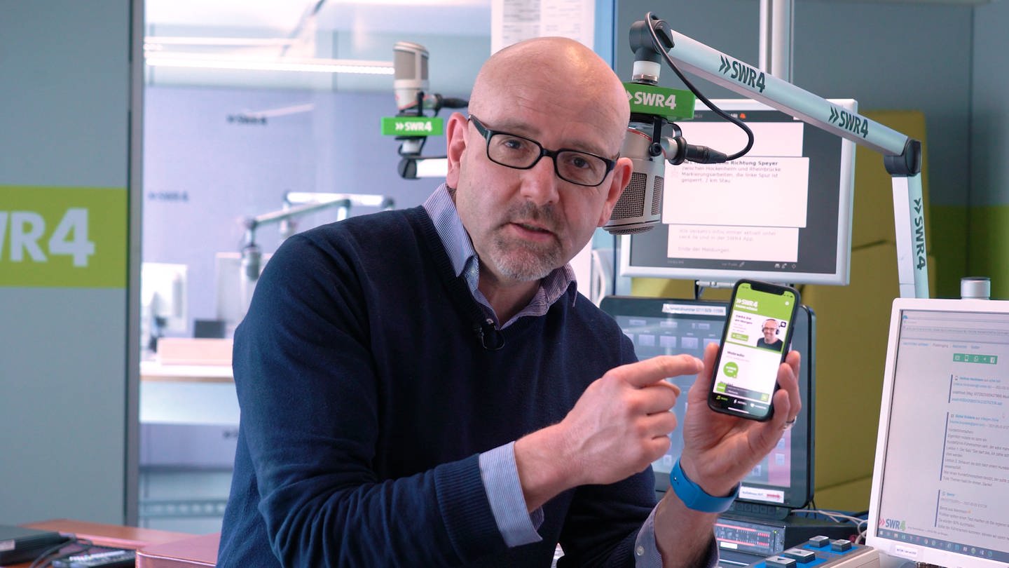 SWR4 Moderator Jörg Assenheimer hält im Hörfunkstudio ein Smartphone in der Hand und erklärt die SWR4 App (Foto: SWR)