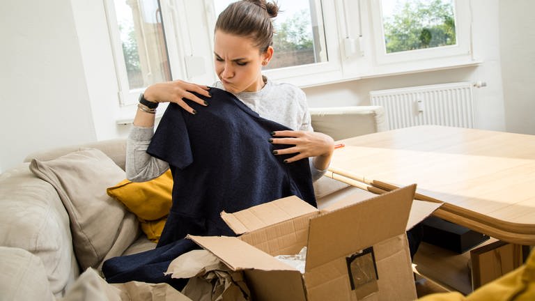  Eine Frau sitzt auf einer Couch und packt ein Päckchen mit einer Kleider-Bestellung aus