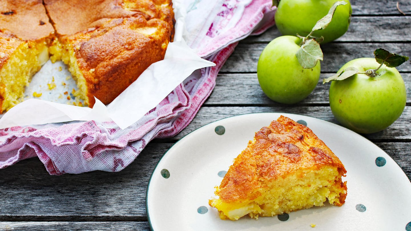 Stück Rührteigkuchen mit Äpfeln auf Teller, daneben ein angeschnittener Kuchen in Backform und Äpfel (Foto: SWR, Imogen Voth)