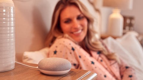Junge Frau sitzt im Bett und lächelt einen Smart Speaker an, der auf dem Nachttisch steht (Foto: IMAGO, Shotshop)