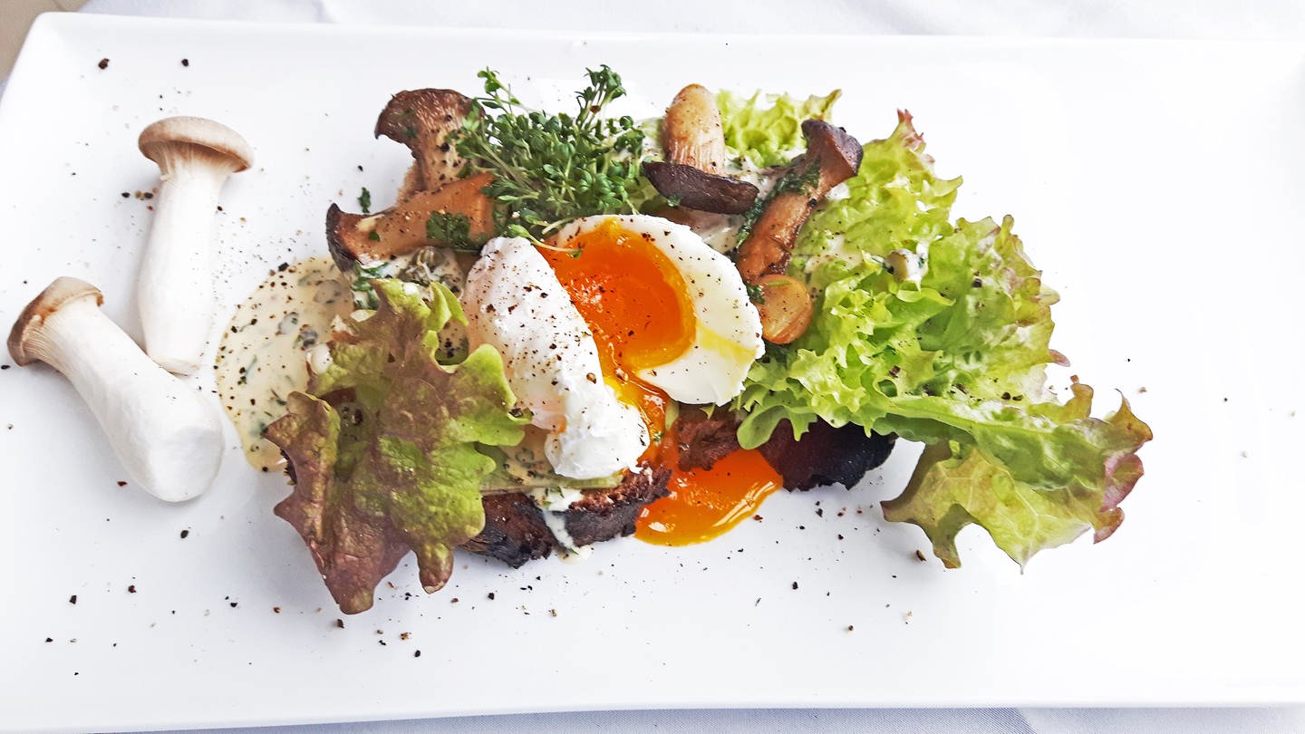 dunkles Brot mit Salat, Pilzen und einem weich gekochten Ei belegt. Daneben zwei Pilze auf dem weißen Teller. (Foto: SWR, Jens Alinia)