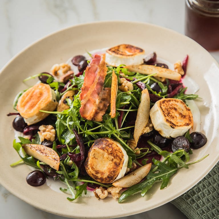 Ziegenkäsetaler auf Salat mit etwas Speck und Trauben, daneben Besteck und  (Foto: dpa Bildfunk, Picture Alliance)