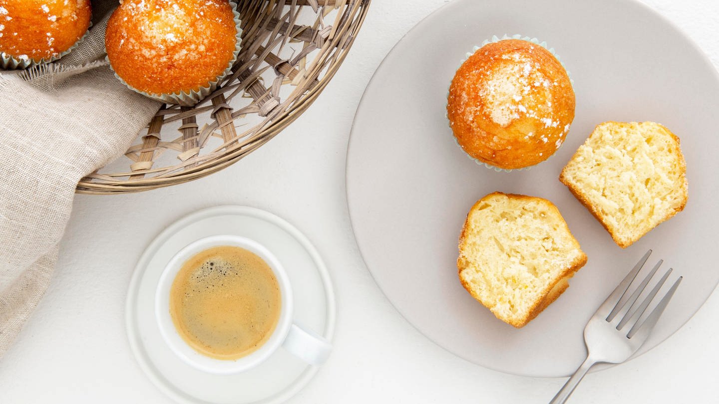 Auf einem Teller sind zwei Zitronenmuffins, einer von beiden ist halbiert. Daneben stehen eine Tasse Kaffee und ein Korb mit weiteren Muffins. (Foto: picture-alliance / Reportdienste, picture alliance / Zoonar | Oleksandr Latkun)