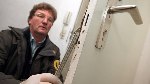 Einbruch – was tun? Ein Polizist begutachtet den Schaden nach einem Einbruch und misst mit einem Zollstock etwas an einer beschädigten Tür nach. (Foto: IMAGO, biky)