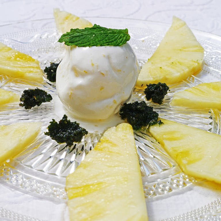 Rezept für ein Dessert: Zitroneneis mit in Rum marinierter Ananas auf einem Glasteller. Auf der Eiskugel in der Mitte liegt ein Minzblatt. (Foto: SWR, Stefanie Kühn)