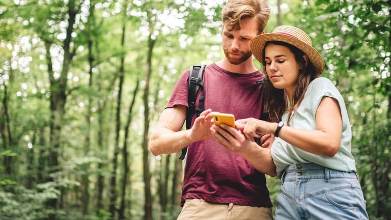 Kostenlose Wander-App: Junger Mann und junge Frau schauen im Wald auf ein Smartphone