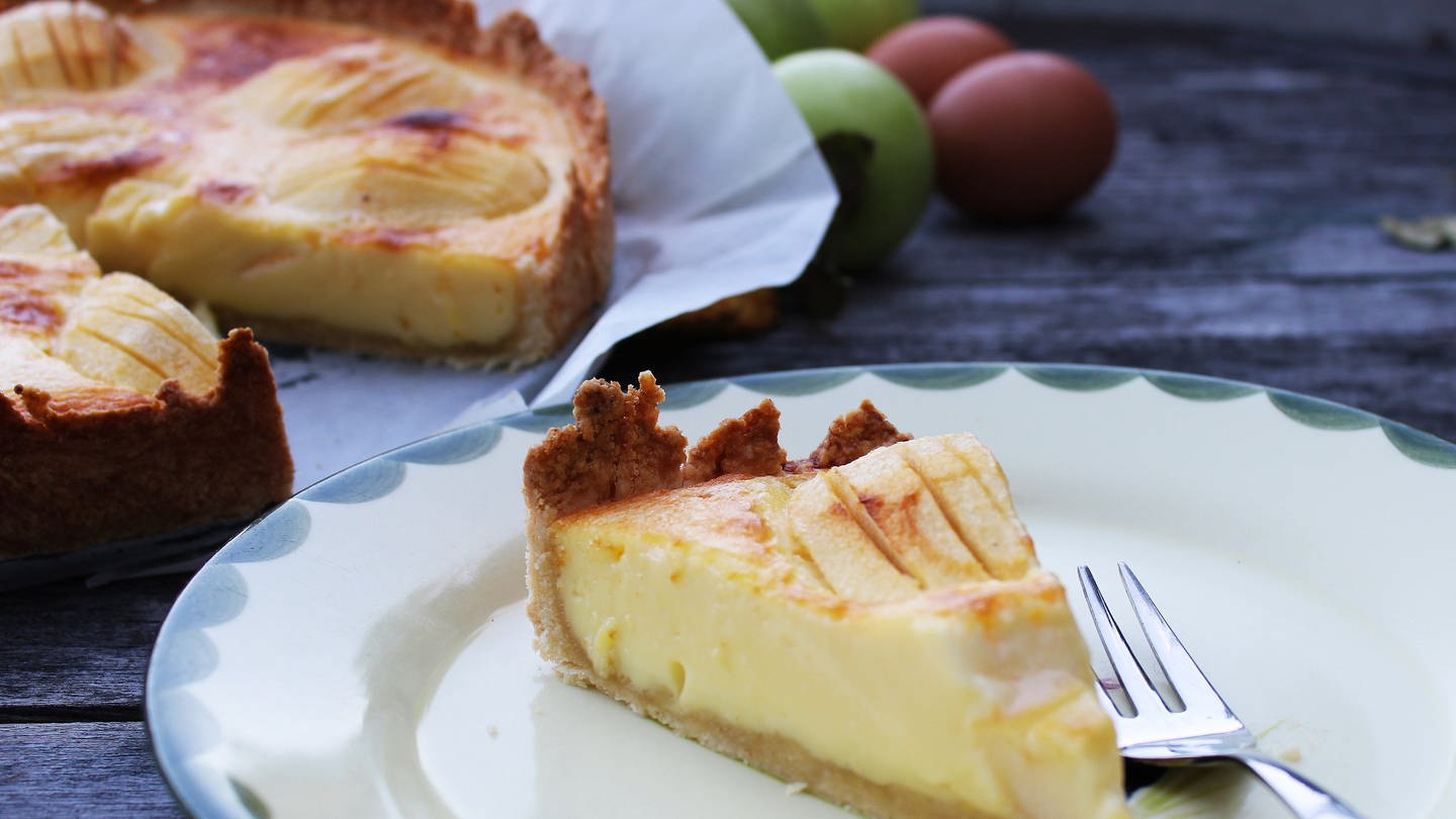 Stück Elsässer Apfelkuchen auf einem Teller, Äpfel und ein angeschnittener Kuchen im Hintergrund (Foto: SWR, Imogen Voth)