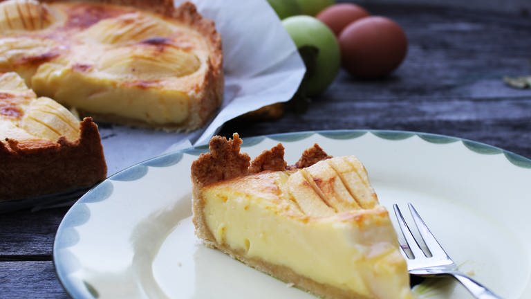 Stück Elsässer Apfelkuchen auf einem Teller, Äpfel und ein angeschnittener Kuchen im Hintergrund (Foto: SWR, Imogen Voth)