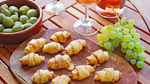 Holzbrett mit Käsehörnchen, daneben Trauben und ein Glas Rose-Wein