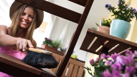 Balkon fit für den Frühling machen: Eine junge Frau macht mit einem Handbesen Gartenmöbel sauber.